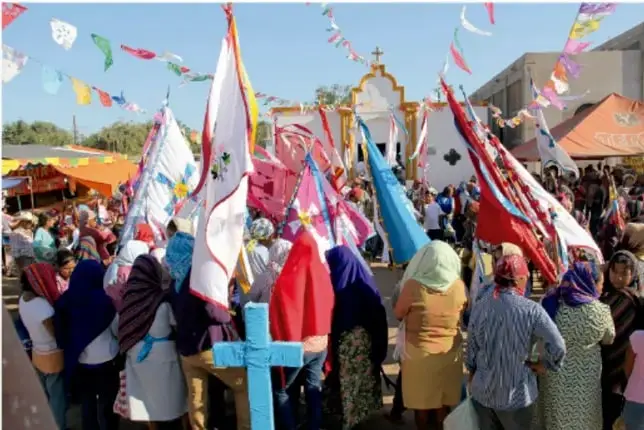 Procesión comunitaria al aire libre con personas portando banderas de colores y una cruz azul, en un ambiente festivo con banderines.