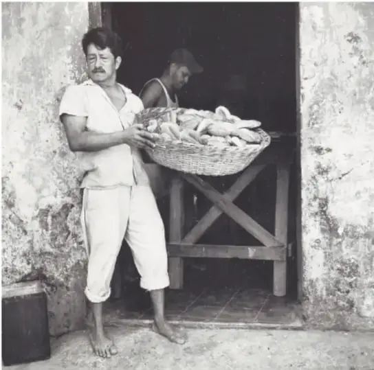 Fotografía en blanco y negro de un panadero descalzo sosteniendo una canasta con pan frente a una puerta, con otra persona al fondo; El pan, 1980, Tlacotalpan, Veracruz, de Mariana Yampolsky (1925-2002).
