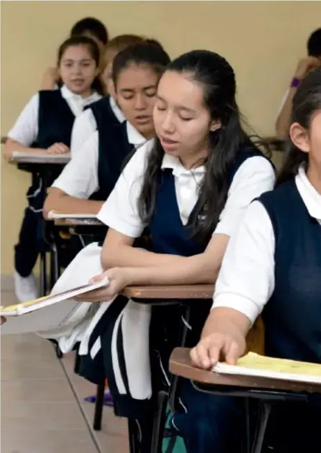 Fotografía de estudiantes de secundaria con uniforme (chamarra y camisa blanca) sentados en sus pupitres en el aula, leyendo y tomando notas.