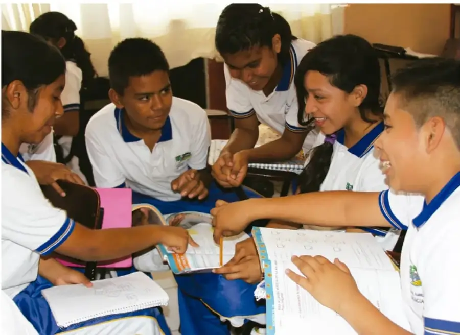 Fotografía de un grupo de estudiantes de telesecundaria con uniforme blanco y azul reunidos alrededor de un escritorio, señalando y comentando libros y cuadernos, participando activamente en una actividad de diálogo en el aula.