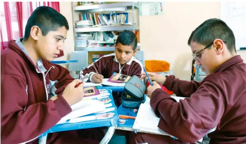 Fotografía de tres estudiantes de telesecundaria trabajando en equipo en un pupitre con cuadernos, lápices y una gorra; están elaborando materiales del proyecto.