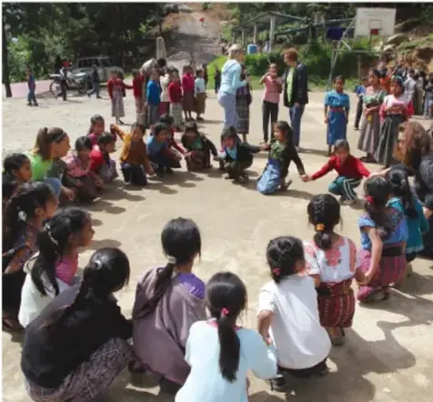 Niñas y niños sentados en círculo en el patio de una escuela, acompañados por adultos, realizando una actividad grupal participativa.
