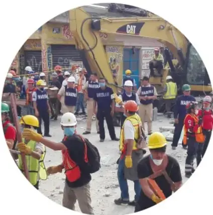 Fotografía circular que muestra a un grupo de personas, algunas con cascos y chalecos reflectantes, reunidas en una calle con escombros y una máquina excavadora amarilla. Parecen ser brigadistas o voluntarios después de un sismo.