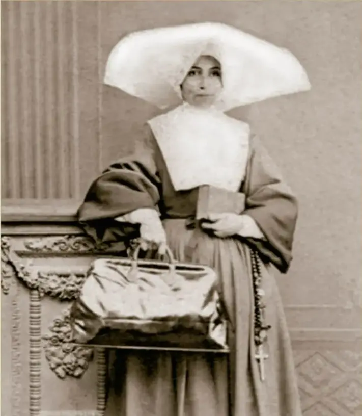 Fotografía sepia titulada 'Monja en estudio fotográfico, tarjeta de visita, ca. 1865' de Becerril: una monja de hábito claro con toca alada sostiene un libro y un rosario junto a una mesa con un cofre. Cortesía INAH/SINAFO.FN.MX.