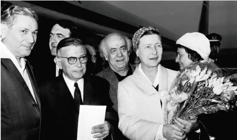 Fotografía en blanco y negro de Simone de Beauvoir de edad madura, con pañuelo en la cabeza y abrigo claro, recibiendo un ramo de flores entre un grupo de personas en lo que parece una llegada de aeropuerto.