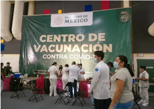 Foto de un Centro de Vacunación contra COVID-19 del Gobierno de México, con personal de salud atendiendo a personas en sillas; cartel verde con letras blancas.