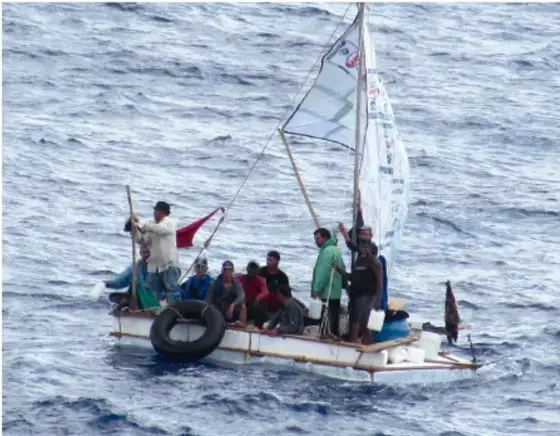Fotografía de un grupo de personas migrantes en una embarcación precaria con una vela blanca y un neumático como flotador, navegando en aguas del mar Mediterráneo.
