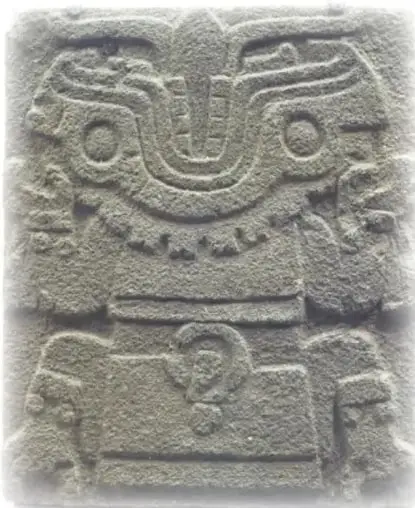 Escultura de piedra tallada en bajo relieve: rostro con tocado de plumas, ojos circulares, collar y elementos ornamentales; pieza prehispánica gris.