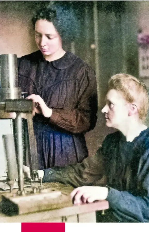 Fotografía antigua en color de Marie Curie de pie junto a su esposo Pierre Curie, quien está sentado trabajando en un aparato científico de laboratorio.