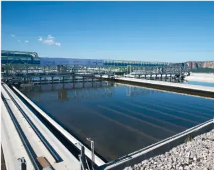 Fotografía de una planta de tratamiento de agua con tanques rectangulares de decantación y estructuras metálicas junto al agua.