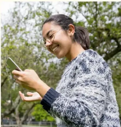 Fotografía de una adolescente sonriente usando un teléfono celular al aire libre, con árboles de fondo.