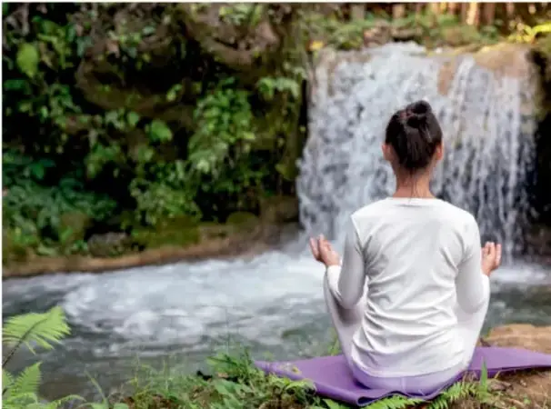 Fotografía de una persona con ropas claras meditando en posición de loto sobre una esterilla, frente a una cascada en un entorno natural verde.