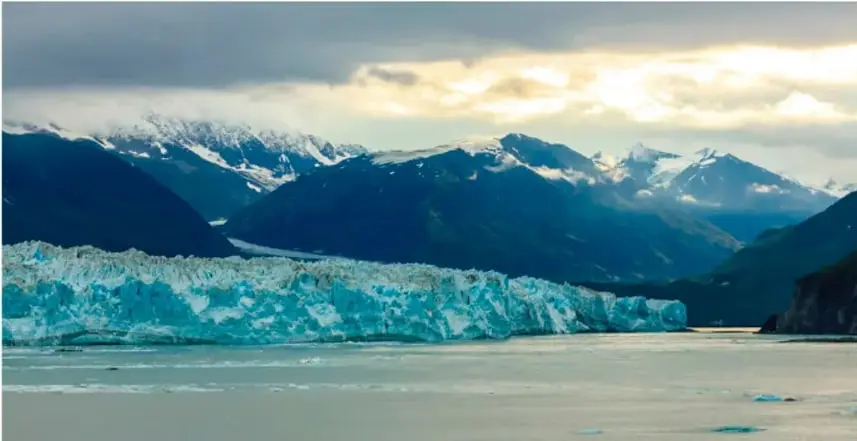 Fotografía de un glaciar azul junto a un lago, con montañas nevadas al fondo bajo un cielo nublado; ilustra la erosión glaciar.