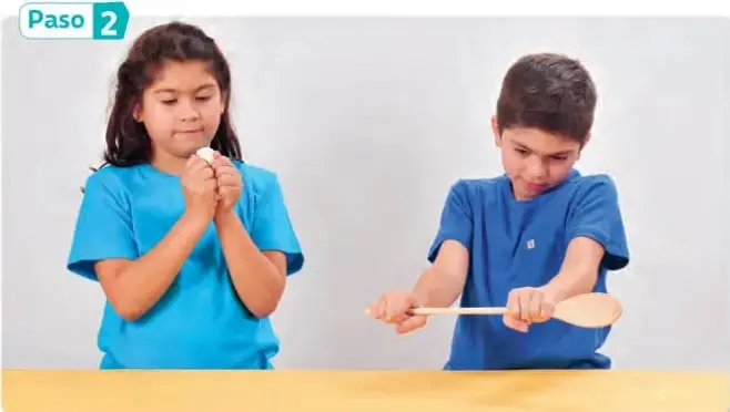 Dos estudiantes en pareja: una niña sostiene y aprieta el guante de goma y un niño sostiene la cuchara de madera sobre una mesa, manipulando los materiales.