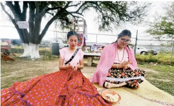 Fotografía de dos mujeres indígenas sentadas al aire libre cosiendo y trabajando textiles tradicionales coloridos bajo un árbol.