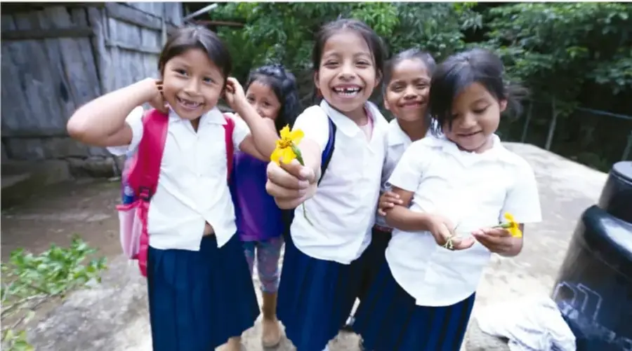 Fotografía de un grupo de niñas con uniforme escolar (camisa blanca y falda azul) sonriendo al aire libre; una de ellas sostiene una flor amarilla. Al fondo se observan árboles y una construcción rústica.