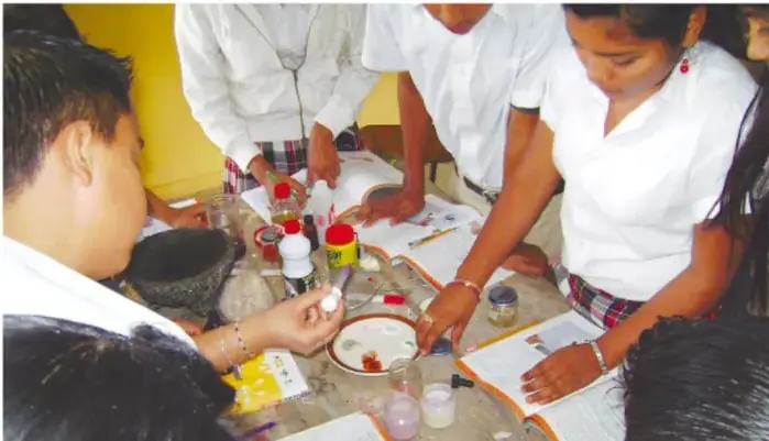 Fotografía de estudiantes de secundaria trabajando en equipo alrededor de una mesa con materiales de laboratorio, libros y cuadernos, realizando una actividad práctica.