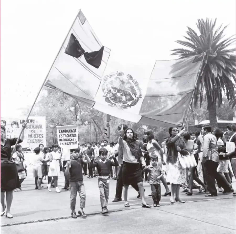 Fotografía en blanco y negro de una marcha o manifestación con la bandera de México al frente; participan mujeres, hombres y niños; se leen pancartas contra la violencia, una de ellas con la frase "La violencia está en contra nosotros, no en nosotros" y otra de una unión nacional de mujeres; al fondo hay palmeras y arbolado urbano.