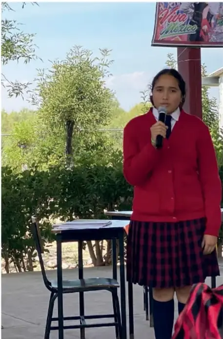 Fotografía de una estudiante de telesecundaria con uniforme (chamarra roja y falda a cuadros) sosteniendo un micrófono, en un acto escolar al aire libre con la leyenda 'Vive México'