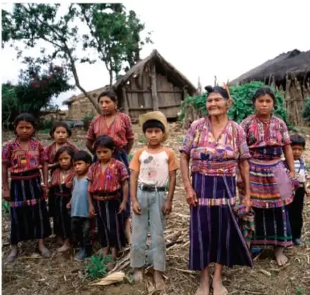 Fotografía de un grupo de personas indígenas, mujeres, hombres y niños con vestimenta tradicional, de pie frente a una vivienda rural de madera y techo de lámina, en un entorno natural.