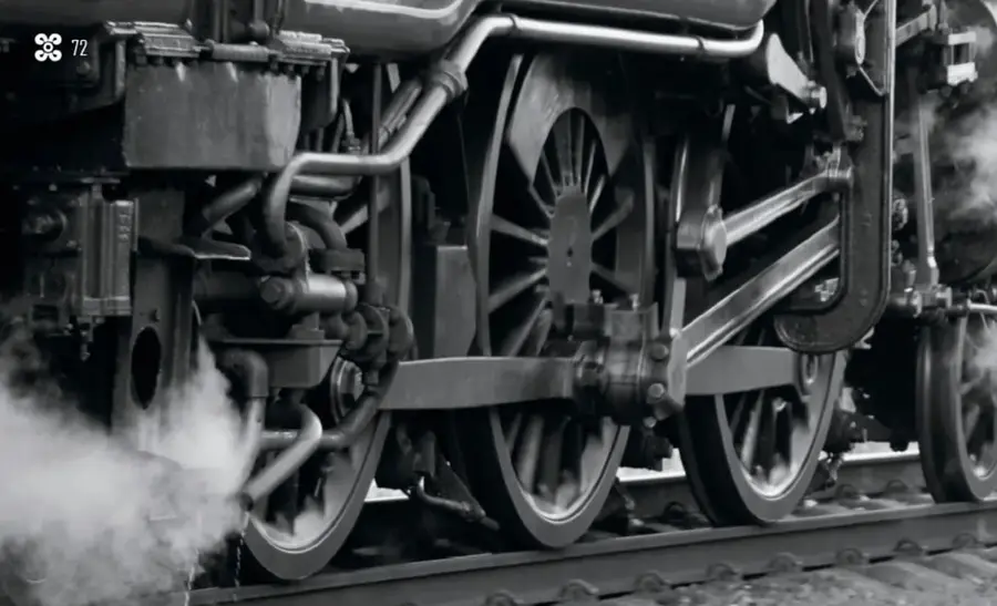 Fotografía en tonos grises de las ruedas y mecanismos de una locomotora de vapor antigua, con humo de vapor saliendo entre la maquinaria mientras avanza sobre las vías.