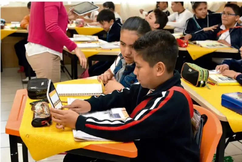 Estudiantes de primaria sentados en sus pupitres usando tabletas y cuadernos; una docente de pie supervisa el trabajo en un aula con mesas amarillas.