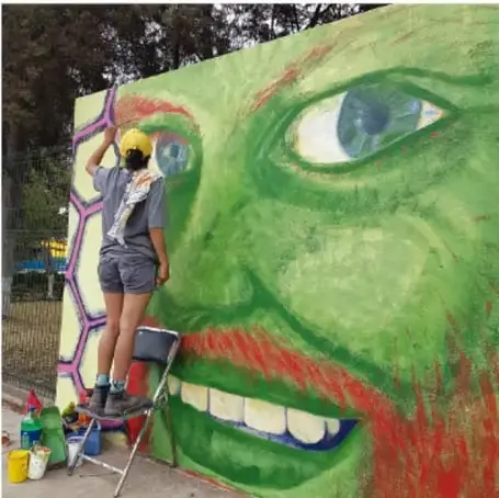 Joven sobre una escalera pintando un mural al aire libre con el rostro verde de un personaje con ojos expresivos y sonrisa (estilo imaginario o fantástico).