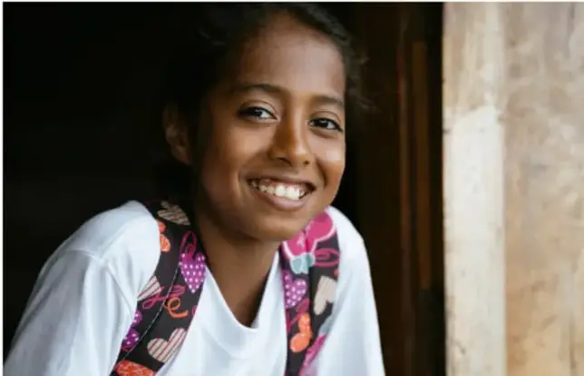 Fotografía de una joven sonriente con mochila de colores, de pie junto a una pared, ilustrando a los jóvenes de las comunidades afromexicanas.