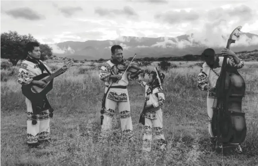 Fotografía en blanco y negro de músicos huicholes con guitarra, violín, contrabajo y una niña, en un campo abierto con montañas al fondo.