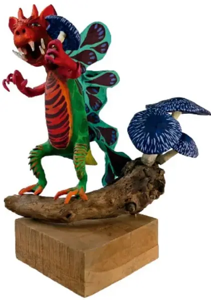 Alebrije de criatura con cabeza roja, alas azules con puntos blancos, cuerpo verde con rayas rojas, montado sobre una rama de madera que descansa en una base de madera.
