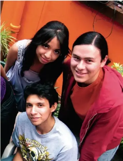Fotografía de tres adolescentes (dos mujeres y un hombre) sonriendo, con fondo de pared naranja. Ilustra la presencia y participación de los NNA.