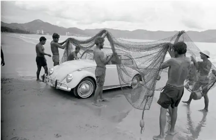 Pescadores extendiendo una red de pesca sobre la playa junto a un Volkswagen blanco. Fotografía en blanco y negro titulada 'Volkswagen con redes en una playa, ca. 1960'.
