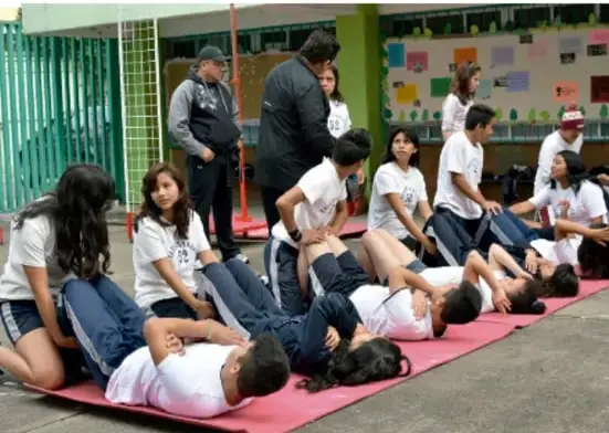 Fotografía de un grupo de estudiantes realizando ejercicios físicos en colchonetas en el patio de una escuela, acompañados por adultos.