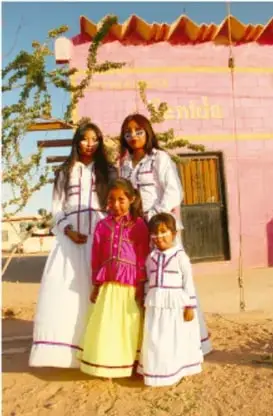 Fotografía de una familia seri frente a una construcción con palmas: una mujer adulta y niñas con vestidos tradicionales largos de colores (blancos, amarillo y magenta) con motivos floreados.