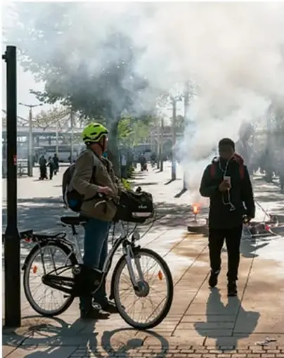 Fotografía de una calle con humo denso, un ciclista con casco y una persona caminando con cubrebocas, reflejando la contaminación del aire.