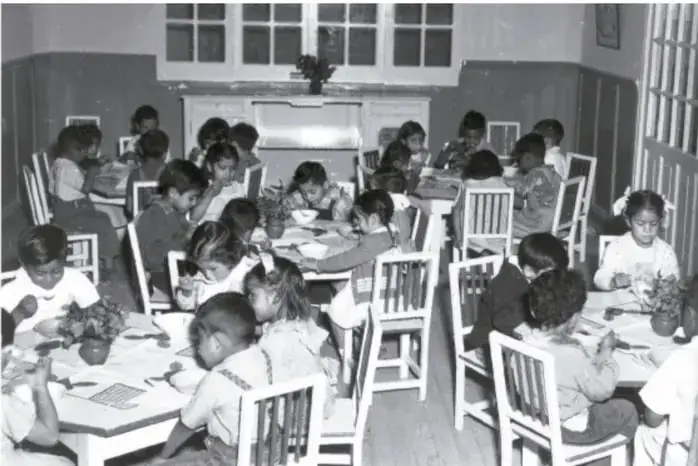 Fotografía en blanco y negro de un salón de clases con muchos estudiantes sentados en pupitres de madera, trabajando en sus cuadernos; al fondo hay ventanas y un pizarrón.