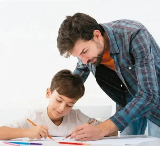 Un adulto (padre o maestro) supervisa a un niño que escribe o dibuja con lápices de colores sobre una mesa.