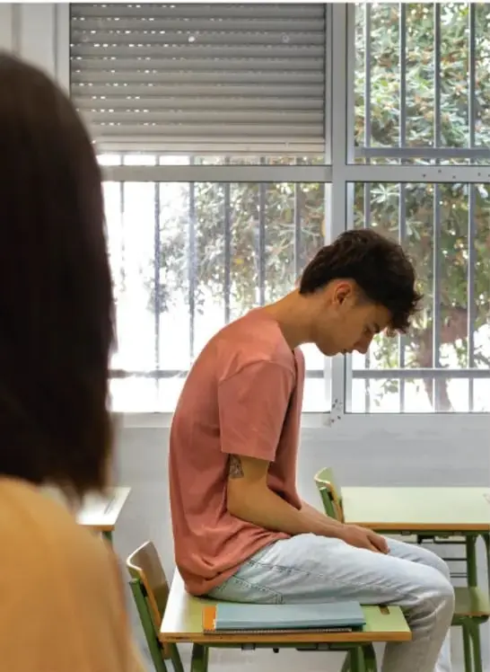 En un salón de clases, un estudiante está sentado con la cabeza gacha y actitud triste mientras una figura femenina de espaldas lo observa, sugiriendo intimidación o violencia psicológica.