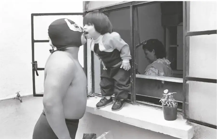 Fotografía en blanco y negro de El Brazo de Plata con máscara, de pie frente a una ventana; un niño se asoma y una mujer observa al fondo.