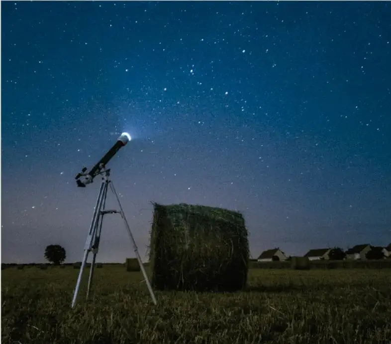 Telescopio sobre trípode apuntando al cielo nocturno estrellado, en un campo con una paca de heno y casas al fondo.