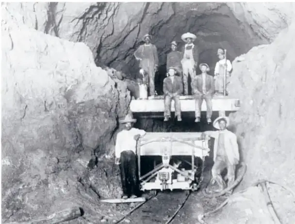 Fotografía histórica en blanco y negro de un grupo de mineros dentro de una mina subterránea, junto a un vagoneta sobre rieles en el interior de la galería.