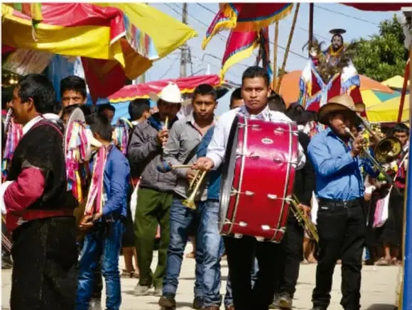 Fotografía de una comparsa o desfile comunitario: músicos con instrumentos de viento y un tambor rojo, portando banderas multicolores, en una calle durante una celebración popular.