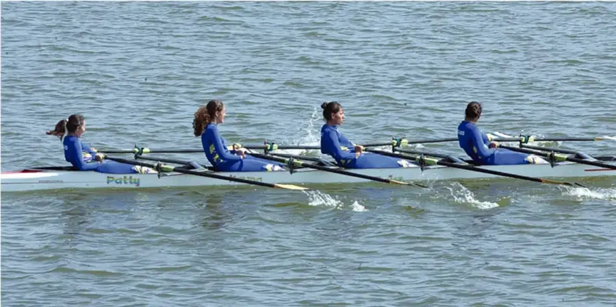 Fotografía de cuatro remeras remando sincronizadas en un bote largo sobre un río, con uniforme azul; metáfora del trabajo en equipo coordinado.