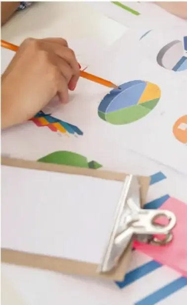 Fotografía de un estudiante trabajando con lápiz sobre hojas con gráficas circulares de colores, un portapapeles con clip metálico y cintas de colores, sobre un escritorio.