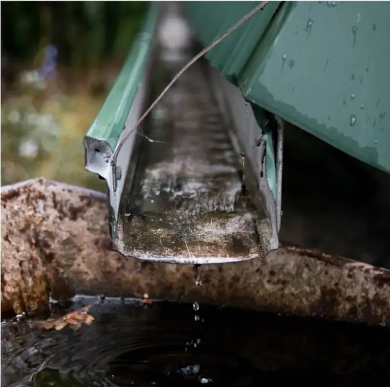 Fotografía de un canal de agua metálico desbordándose y cayendo en gotas hacia una alberca o recipiente de agua, sobre una base de concreto.