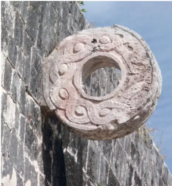 Anillo de piedra tallado con motivos curvos (marcador o aro de juego de pelota) colocado en un muro de piedra de una zona arqueológica.