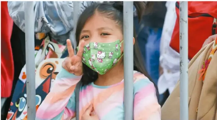 Fotografía de una niña con cubrebocas que hace el gesto de victoria tras una reja, expresando alegría.