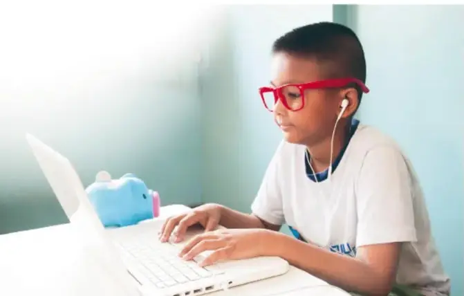 Niño con lentes rojos y audífonos usando una laptop, sentado en un escritorio.
