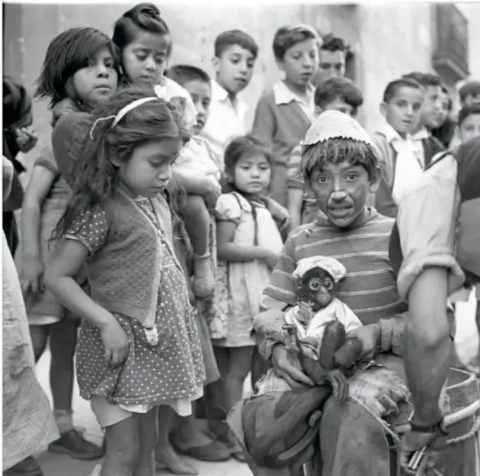 Niño disfrazado de payasito con un mono de juguete, rodeado de otros niños que lo observan. Fotografía en blanco y negro titulada 'Payasito con mono, escena fellinesca, ca. 1950'.