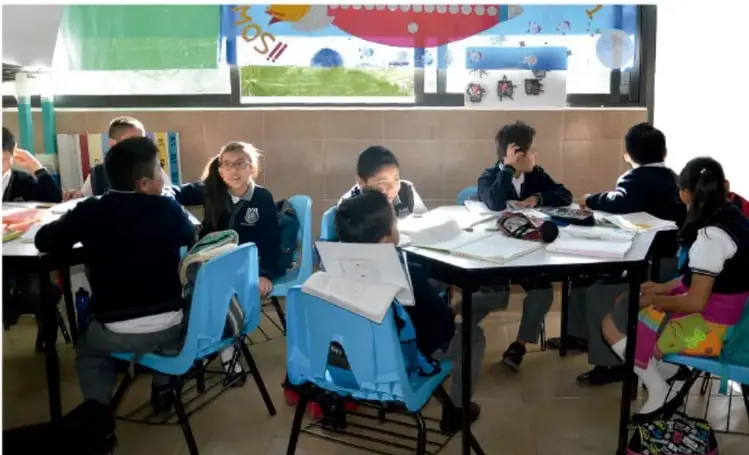 Fotografía de un grupo de estudiantes de primaria sentados en mesas azules dentro de un aula, trabajando en equipo con cuadernos y libros; al fondo hay un mural colorido.